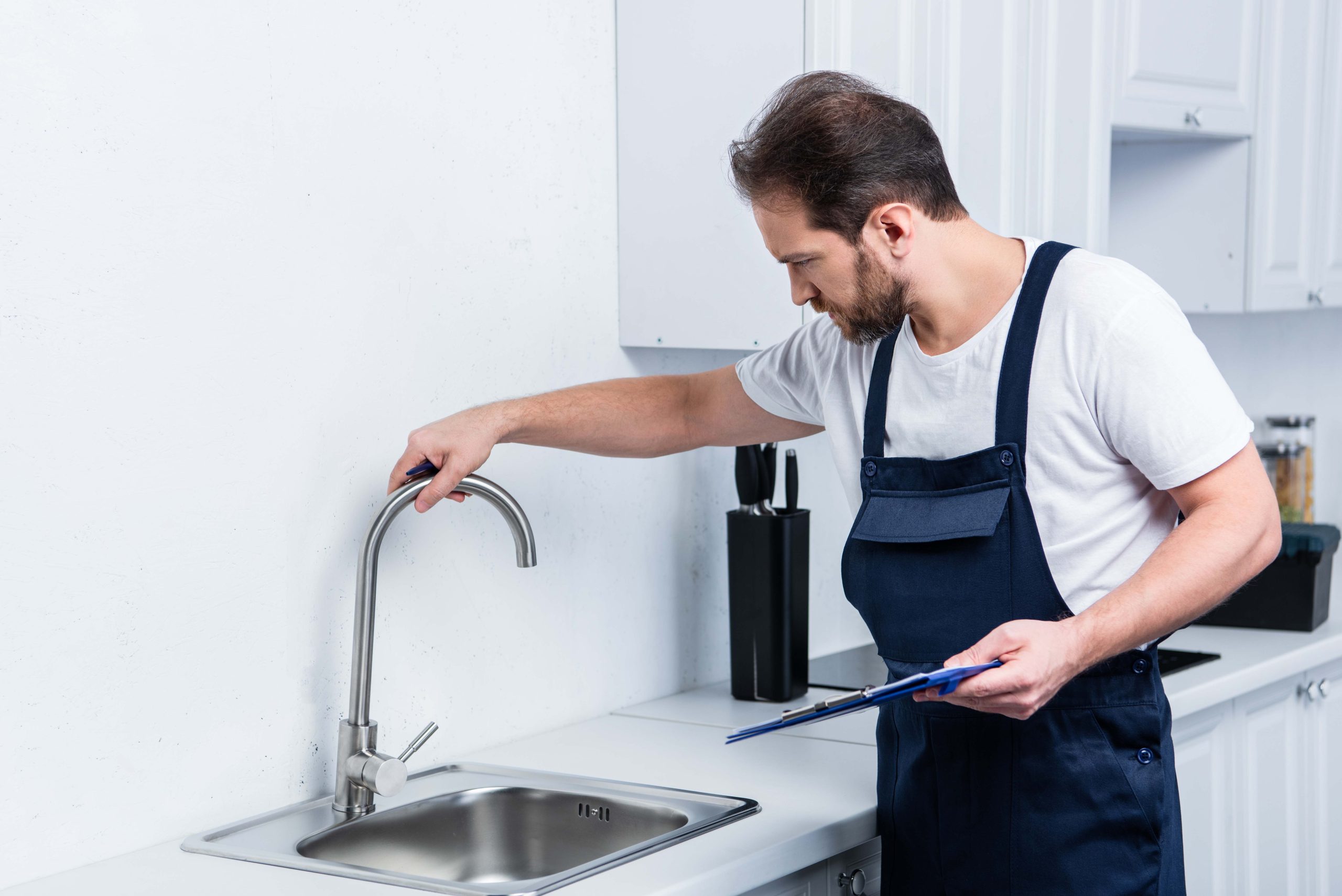 repairman in working overall holding clipboard and checking sink